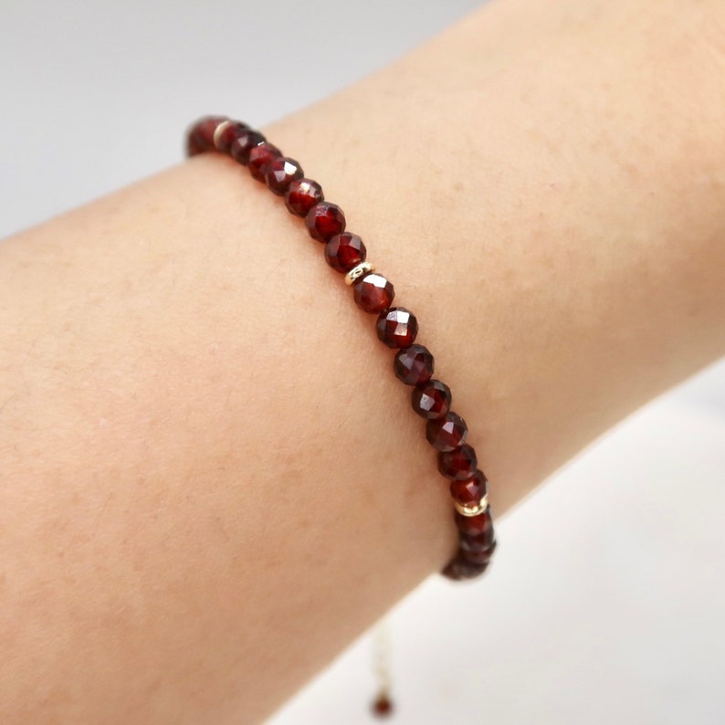 Pu&ograve; includere: Un delicato braccialetto con perline di granato rosso intenso sfaccettate. Le perline sono infilate con piccoli distanziatori color oro. Il braccialetto &egrave; mostrato su un polso, evidenziando il design e il colore del gioiello.