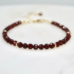 Pu&ograve; includere: Un delicato braccialetto con piccole perline di granato rosso scuro sfaccettate infilate insieme. Il braccialetto include piccole perline distanziatrici color oro e una catena regolabile color oro. Il braccialetto &egrave; esposto su una superficie bianca.