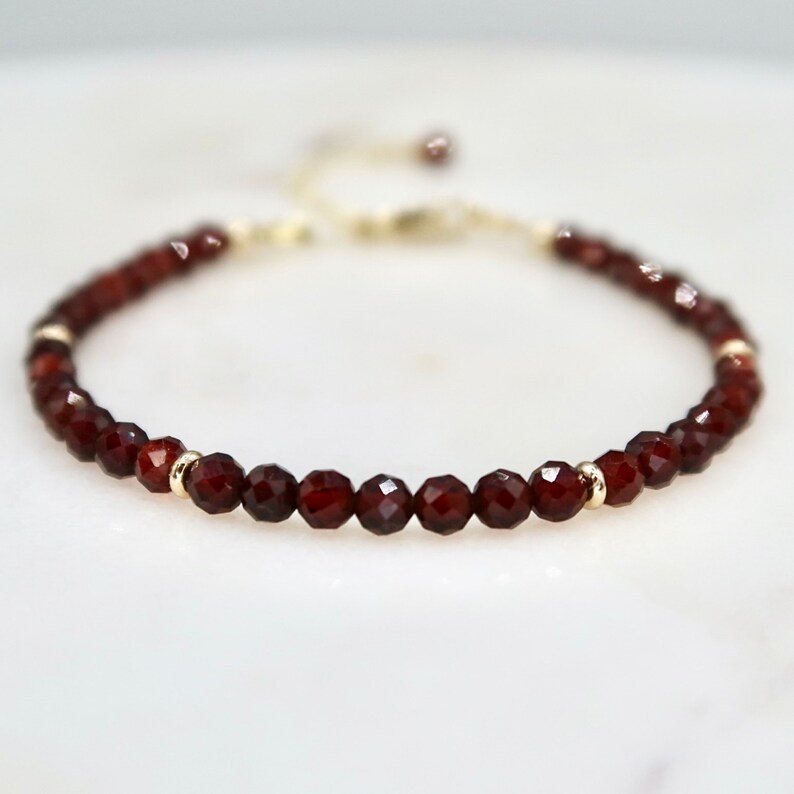 Pu&ograve; includere: Un delicato braccialetto con piccole perline di granato rosso scuro sfaccettate infilate insieme. Il braccialetto include piccole perline distanziatrici color oro e una catena regolabile color oro. Il braccialetto &egrave; esposto su una superficie bianca.