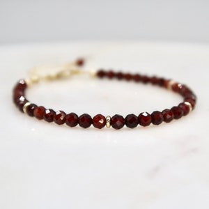 Pu&ograve; includere: Un delicato braccialetto con perline di granato rosso intenso sfaccettate. Le perline sono infilate con una catena color oro e piccoli accenti dorati. Il braccialetto &egrave; regolabile.