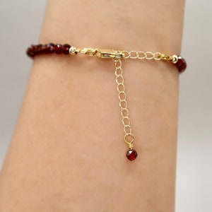 Pu&ograve; includere: Un delicato braccialetto con piccole perline rotonde di colore rosso scuro. Il braccialetto ha una chiusura color oro e una catena regolabile. Una singola perla rossa pende dall'estremit&agrave; della catena. Il braccialetto &egrave; mostrato su un braccio.