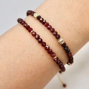 Pu&ograve; includere: Due braccialetti di perline con piccole perline sfaccettate rosso scuro. I braccialetti hanno dettagli color oro e sono indossati su un polso. Un braccialetto ha un dettaglio a forma di corona. I braccialetti sono infilati su un cordoncino sottile di colore chiaro.