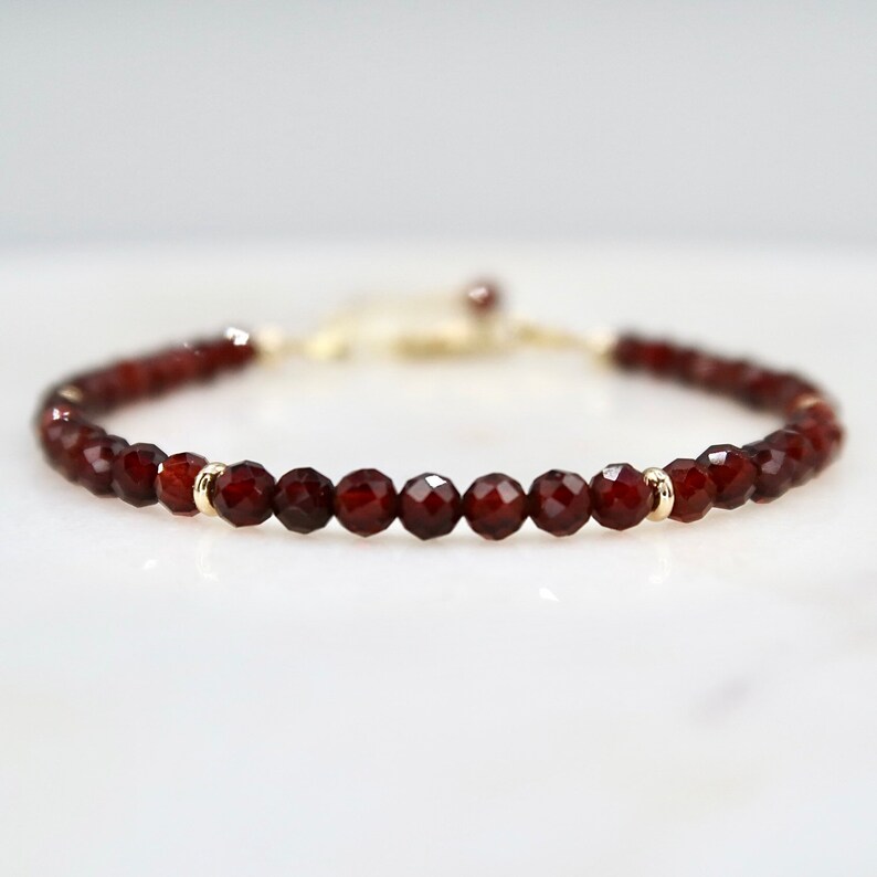 Pu&ograve; includere: Un delicato braccialetto con piccole perline di granato rosso scuro sfaccettate. Le perline sono infilate con dettagli color oro. Il braccialetto &egrave; esposto su una superficie bianca, mettendo in risalto il suo design elegante e il colore.