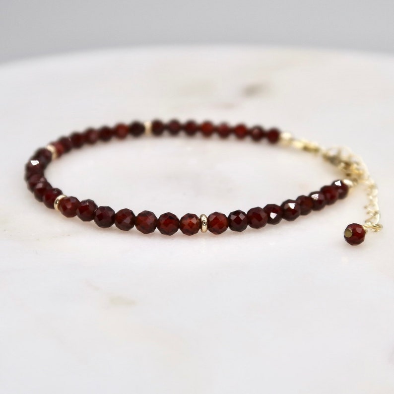 Pu&ograve; includere: Un delicato braccialetto con piccole perline di granato rosso scuro sfaccettate e infilate. Il braccialetto ha dettagli color oro e una catena regolabile abbinata con una singola perla di granato all'estremit&agrave;. Il gioiello &egrave; esposto su una superficie bianca.