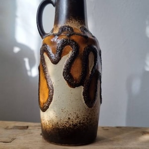 Może przedstawiać: Ceramiczny wazon z ciemnobrązowym uchwytem i obramowaniem, z kremowym korpusem z brązowymi, kroplowymi akcentami. Wazon stoi na zwietrzałej drewnianej powierzchni, prezentując estetykę vintage.