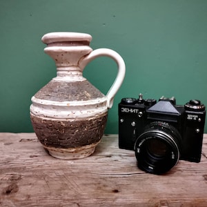 Może przedstawiać: Czarny, zabytkowy aparat fotograficzny z obiektywem i napisem "ЗЕНИТ" obok białego i brązowego ceramicznego dzbanka z uchwytem. Dzbanek ma teksturowany, przypominający korę wzór pośrodku.