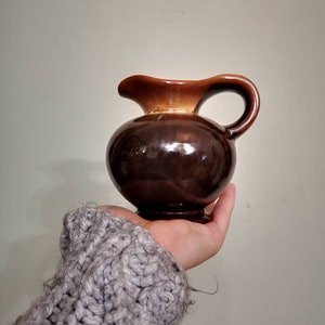 Może przedstawiać: Mały, brązowy ceramiczny dzbanek o zaokrąglonym korpusie i ciemnobrązowym uchwycie. Górna część dzbanka ma jaśniejszy brązowy kolor. Dzbanek jest trzymany w dłoni osoby.