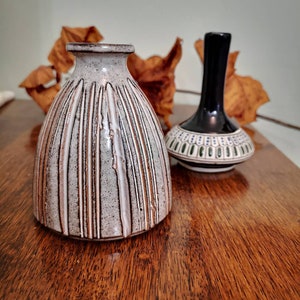 Może przedstawiać: Dwa ceramiczne wazony na drewnianej powierzchni. Większy wazon jest szary z pionowymi brązowymi i białymi paskami. Mniejszy wazon jest czarny z biało-zielonym wzorem na podstawie. Suche liście w tle.