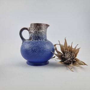 Peut inclure: Un petit pichet en céramique bleu avec une anse et un bec verseur. Le haut du pichet est marron, passant au bleu. Une fleur séchée et brune est à côté du pichet. Le pichet a un corps rond et une petite base.