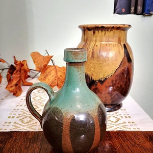 Może przedstawiać: Dwa ceramiczne wazony. Przedni wazon jest turkusowy i brązowy z uchwytem. Tylny wazon jest brązowy i beżowy. Oba wazony mają błyszczące wykończenie i stoją na drewnianej powierzchni.