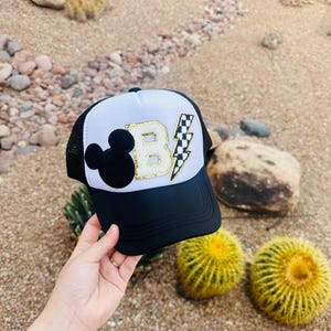 Peut inclure: Casquette de camionneur blanche et noire avec une tête de Mickey Mouse noire, une lettre "B" en paillettes dorées et un éclair noir et blanc à carreaux.
