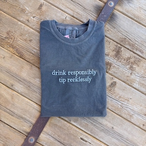 May include: A dark gray t-shirt with the text "drink responsibly tip recklessly" in white script. The shirt is folded and lies on a wooden surface with a metal accent.
