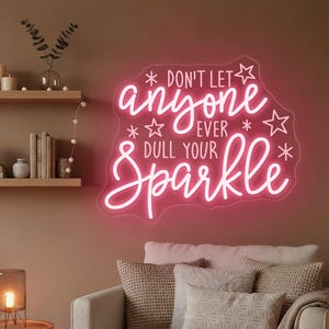 Op de afbeelding: Een roze neonbord met de tekst "DON'T LET ANYONE EVER DULL YOUR SPARKLE" in een sierlijk lettertype. Het bord is bevestigd aan een bruine muur boven een beige bank met kussens en een deken. Links staat een kleine tafel met een lamp.