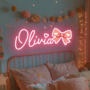 May include: A pink neon sign that spells "Olivia" in cursive with a bow design. The sign is mounted above a bed with pink bedding and pillows. The room is decorated with pink and gold accents.