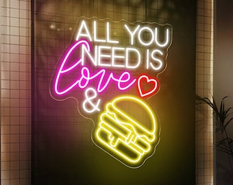 May include: Neon sign with the text "All you need is love &" and a yellow neon burger. The sign is mounted on a black wall with white tile behind it.