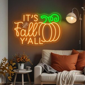 Puede incluir: Un letrero de neón naranja con las palabras "It's Fall Y'all" y un diseño de calabaza verde. El letrero está montado en una pared oscura sobre un sofá blanco con cojines naranjas y una manta. Las calabazas y la decoración otoñal están dispuestas en la habitación.