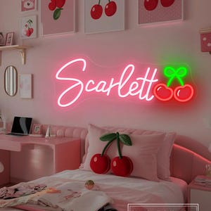 May include: A pink bedroom featuring a neon sign spelling "Scarlett" in cursive with a cherry design. The room includes a pink desk, bed, and cherry-themed decor, such as framed cherry art and cherry pillows.