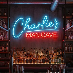 Puede incluir: Un letrero de neón montado en la pared con el texto Charlie's en letra azul y MAN CAVE en letras rojas de molde. El letrero está colocado sobre un estante de bar lleno de varias botellas de licor de vidrio y cristalería.
