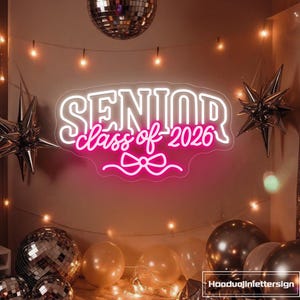 Puede incluir: Un letrero de neón blanco con la palabra "SENIOR" y "class of 2026" en rosa. Decorado con un lazo y globos en forma de estrella. Las bolas de discoteca y las luces de cadena crean un ambiente festivo.