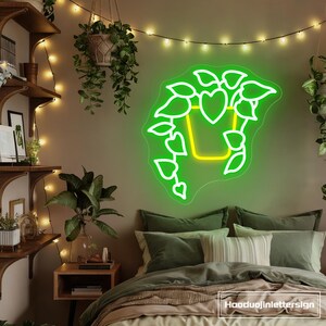 Könnte beinhalten: Ein Neonschild in Form einer Topfpflanze mit grünen Konturen und einem gelben Topf. Das Schild ist an einer beigen Wand über einem Bett mit grüner und beiger Bettwäsche angebracht. Der Raum ist mit Hängepflanzen und Lichterketten dekoriert.