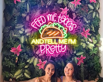 May include: Two people pose in front of a neon sign that reads "Feed me tacos and tell me I'm pretty". The sign features a pink neon taco with green lettuce and yellow cheese. The sign is mounted on a green wall with artificial flowers.