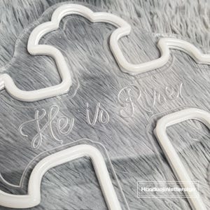 Puede incluir: Un letrero transparente de acr&iacute;lico con forma de nube con las palabras "He is Risen" en una fuente de escritura delicada. El letrero tiene un contorno de ne&oacute;n blanco y est&aacute; sobre un fondo gris suave.