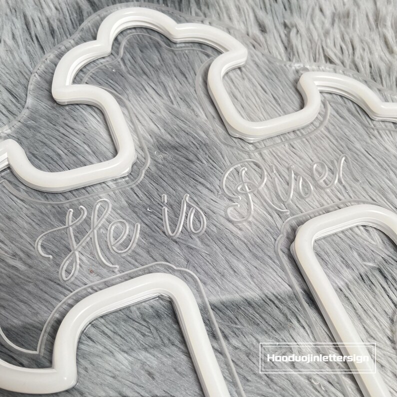 Puede incluir: Un letrero transparente de acr&iacute;lico con forma de nube con las palabras "He is Risen" en una fuente de escritura delicada. El letrero tiene un contorno de ne&oacute;n blanco y est&aacute; sobre un fondo gris suave.