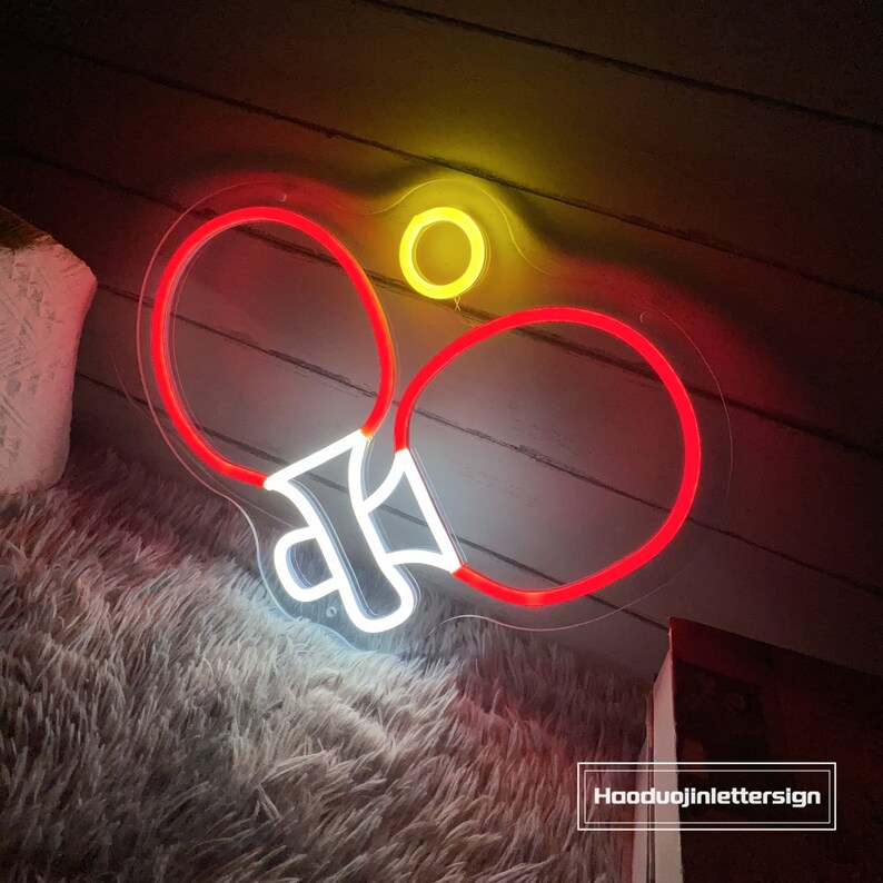 May include: Neon sign depicting two red ping pong paddles and a yellow ball. The sign is mounted on a clear acrylic backing.