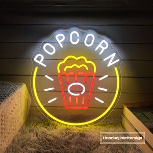 May include: A yellow and white neon sign with the word "POPCORN" in a circle. Inside the circle is a red and yellow neon image of a popcorn bucket.
