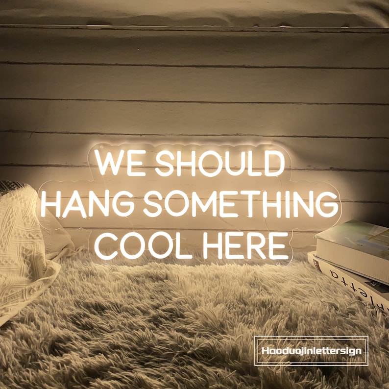May include: A white neon sign with the text "WE SHOULD HANG SOMETHING COOL HERE". The sign is on a light-colored wooden wall, with a fluffy rug and a blanket in the background. The sign is from Hooduojinlettersign.
