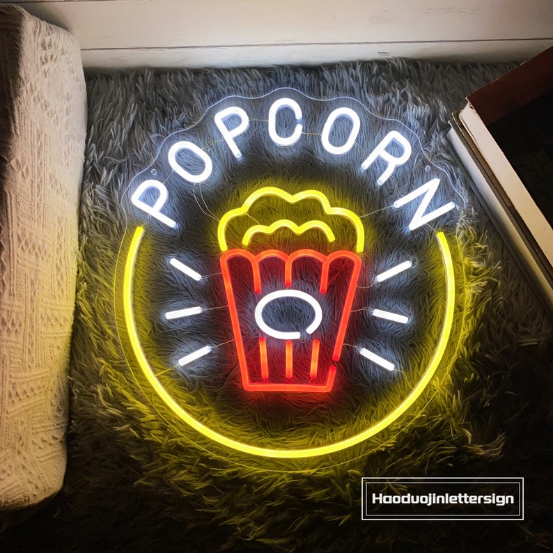 May include: A neon sign with the word "POPCORN" in white letters with a yellow outline. The sign also features a red and yellow popcorn bucket with white outlines.