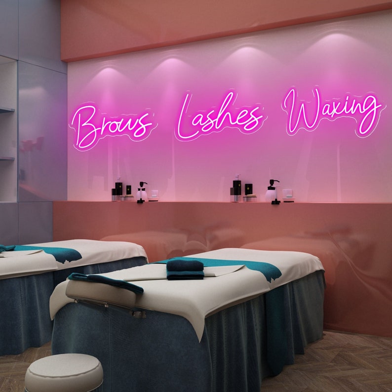 Puede incluir: Un letrero de ne&oacute;n rosa con las palabras "Brows, Lashes, Waxing" en una pared blanca sobre dos mesas de masaje blancas con s&aacute;banas azules. La habitaci&oacute;n tiene un esquema de color rosa y blanco.