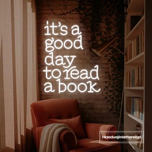 May include: A neon sign with the text "it's a good day to read a book" in white. The sign is mounted on a brick wall. A red armchair, a pillow, and a blanket are in front of the sign. A bookshelf is to the right.