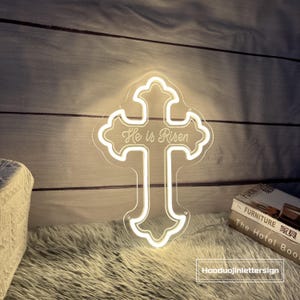 Puede incluir: Una cruz de ne&oacute;n blanca con la frase "He is Risen" en escritura cursiva. La cruz est&aacute; iluminada y montada en una pared de madera. Es un objeto decorativo, ideal para celebraciones religiosas.