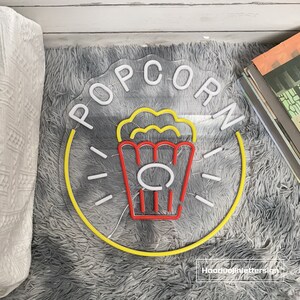 May include: A neon sign with the word "POPCORN" in white letters on a yellow circle. A red and yellow popcorn bucket with popcorn is in the center of the circle.