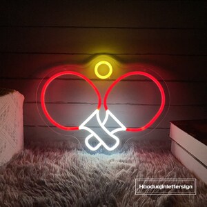 May include: A neon sign depicting two ping pong paddles and a ball in red, white, and yellow. The sign is illuminated and mounted on a wall.