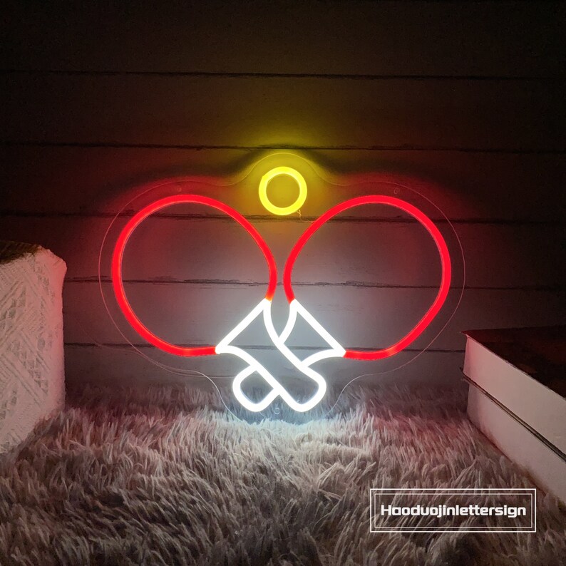 May include: A neon sign depicting two ping pong paddles and a ball in red, white, and yellow. The sign is illuminated and mounted on a wall.