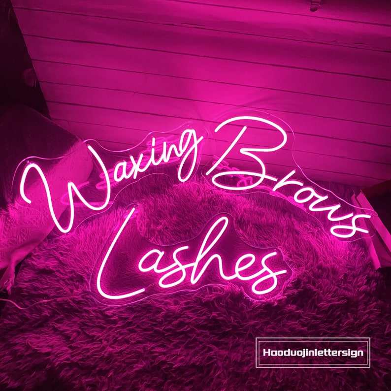 Puede incluir: Letrero de ne&oacute;n rosa con el texto "Waxing Brows Lashes" en una fuente cursiva. El letrero est&aacute; sobre una superficie rosa y peluda.