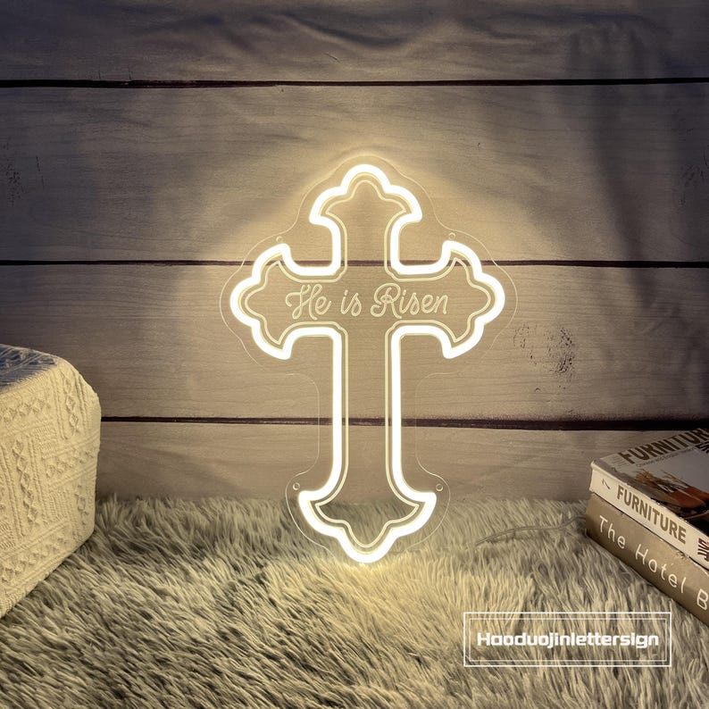 Puede incluir: Un letrero de ne&oacute;n con forma de cruz con las palabras "He is Risen" en una fuente decorativa. El letrero est&aacute; iluminado con una luz blanca c&aacute;lida y est&aacute; montado sobre un respaldo transparente. Es una decoraci&oacute;n religiosa.