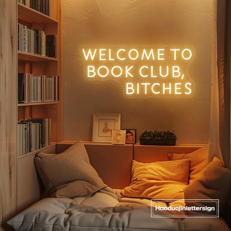 May include: A neon sign that reads "WELCOME TO BOOK CLUB, BITCHES" is mounted on a wall above a cozy reading nook. The nook features pillows and a built-in bookshelf filled with books. The sign and the room are illuminated with warm lighting.