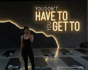 May include: A neon sign with the words "YOU DON'T HAVE TO YOU GET TO" in a gym setting. The sign is illuminated in a warm, inviting glow. The image also shows yoga mats and exercise balls.