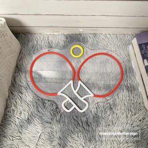 May include: A neon sign depicting two red ping pong paddles with a white ball in the middle. The sign is on a white fluffy surface. The sign has a yellow circle at the top.
