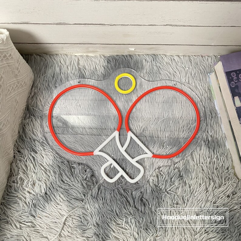 May include: A neon sign depicting two red ping pong paddles with a white ball in the middle. The sign is on a white fluffy surface. The sign has a yellow circle at the top.