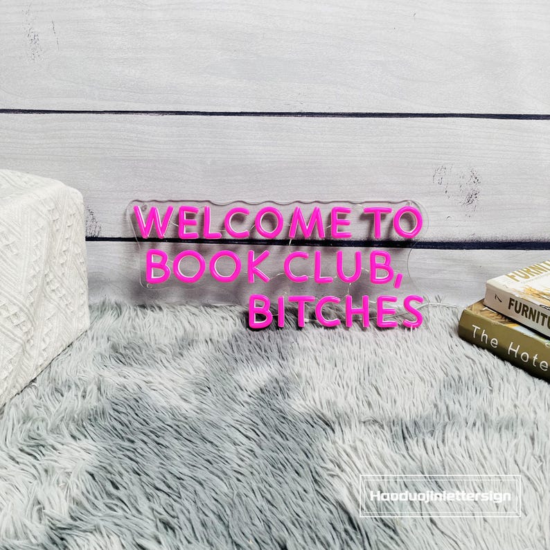 May include: A neon pink sign that reads "WELCOME TO BOOK CLUB, BITCHES" on a clear backing. The sign is placed on a gray fluffy rug in front of a white wooden background. Two books are visible on the right.