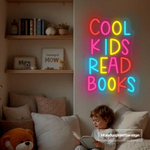May include: A neon sign with the words "COOL KIDS READ BOOKS" in bright pink, yellow, and blue. The sign is mounted on a wall above a child reading a book. The room has bookshelves and pillows.