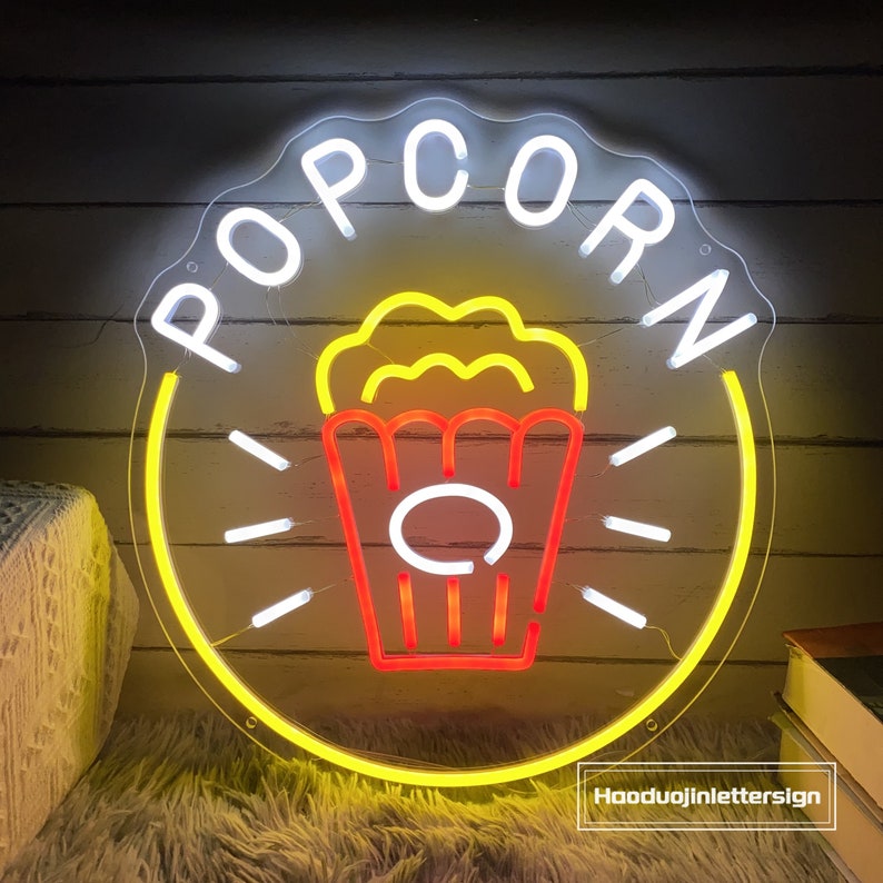 May include: A neon sign with the word "POPCORN" in white letters and a yellow outline. The sign also features a red and yellow popcorn bucket with popcorn spilling out of the top.