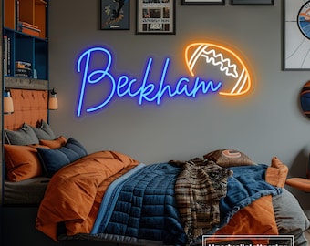 Op de afbeelding: Een blauw neonbord met de naam "Beckham" in een cursief schrift, met daar naast een oranje neon voetbal. Het bord is gemonteerd op een grijze muur boven een bed met oranje en blauwe beddengoed.