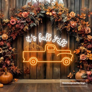Pode incluir: Letreiro de néon laranja e branco com a frase "it's fall y'all" e um camião com abóboras. O letreiro está sobre um fundo de madeira, decorado com folhas de outono e abóboras.