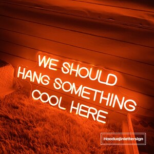 May include: An orange neon sign with the text "WE SHOULD HANG SOMETHING COOL HERE". The sign is illuminated and mounted on a wooden wall. The sign is above a fluffy, orange rug.