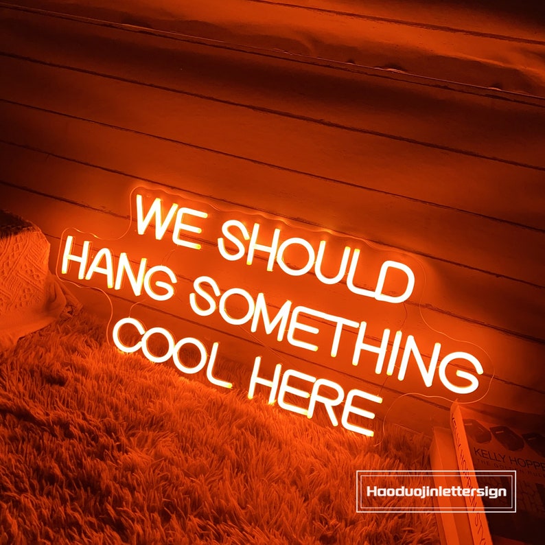 May include: An orange neon sign with the text "WE SHOULD HANG SOMETHING COOL HERE". The sign is illuminated and mounted on a wooden wall. The sign is above a fluffy, orange rug.